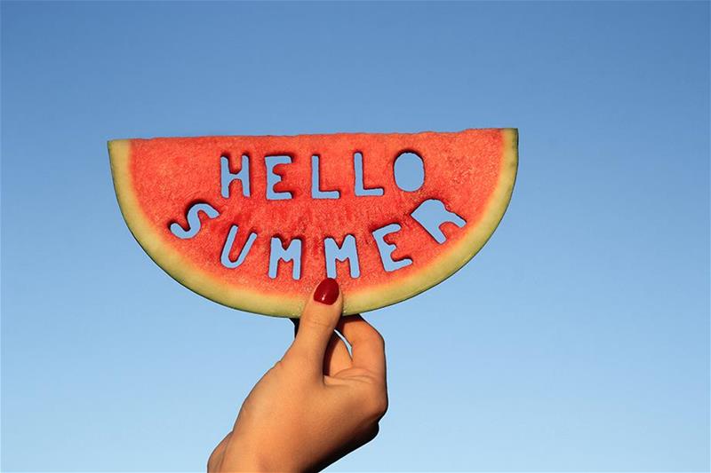 An individual holding a slice of watermelon with the words 'Hello Summer' cut out of it