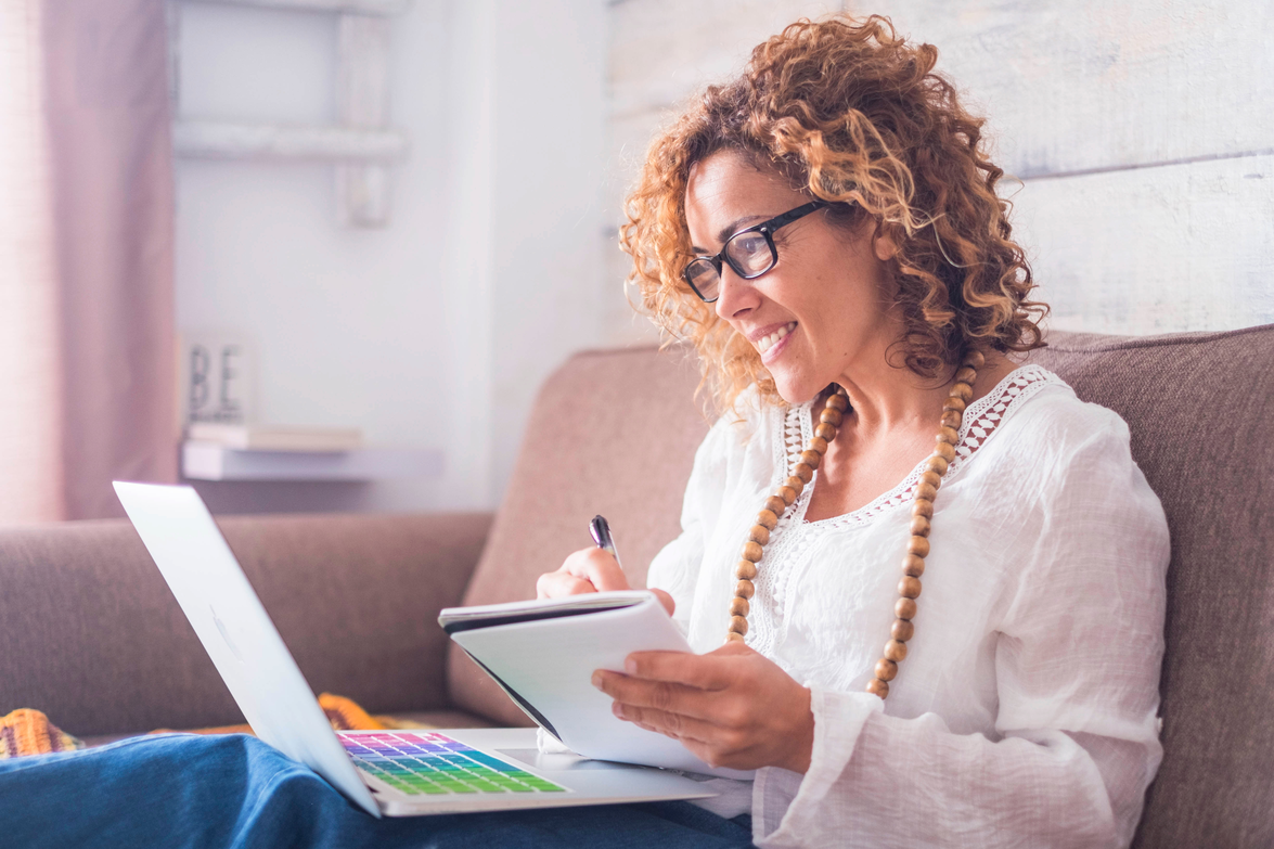 A woman sitting on a couch looking at a laptop and taking notes