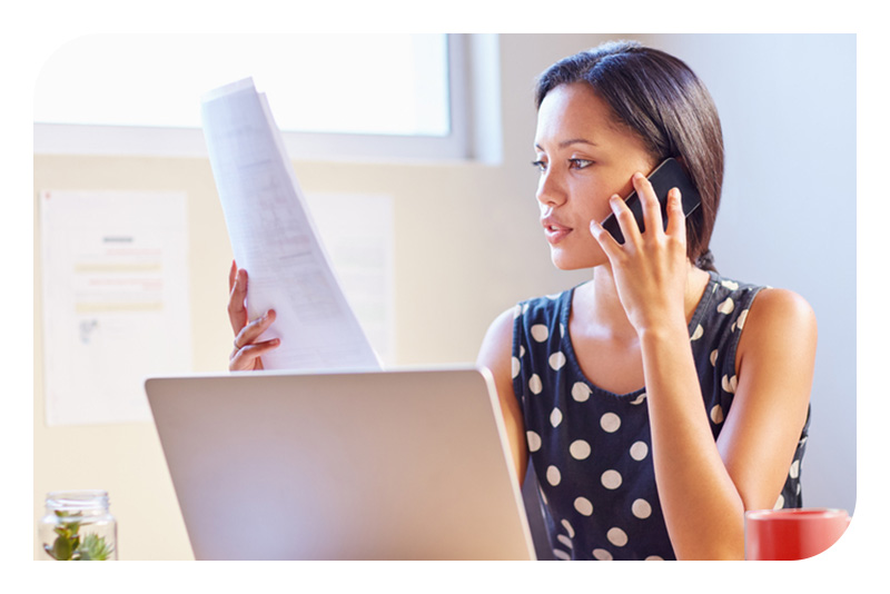 A woman at a desk using a phone while reviewing papers