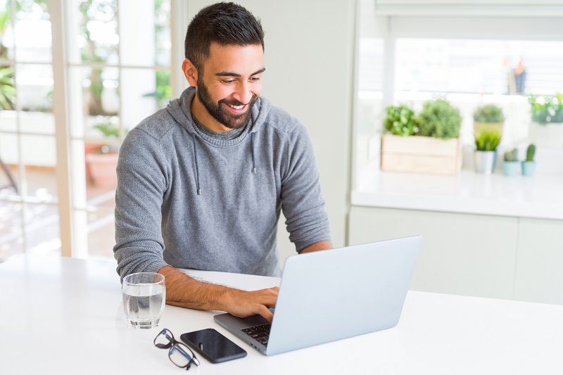 A man in a kitchen using a laptop with water, glasses and a smartphone on the table