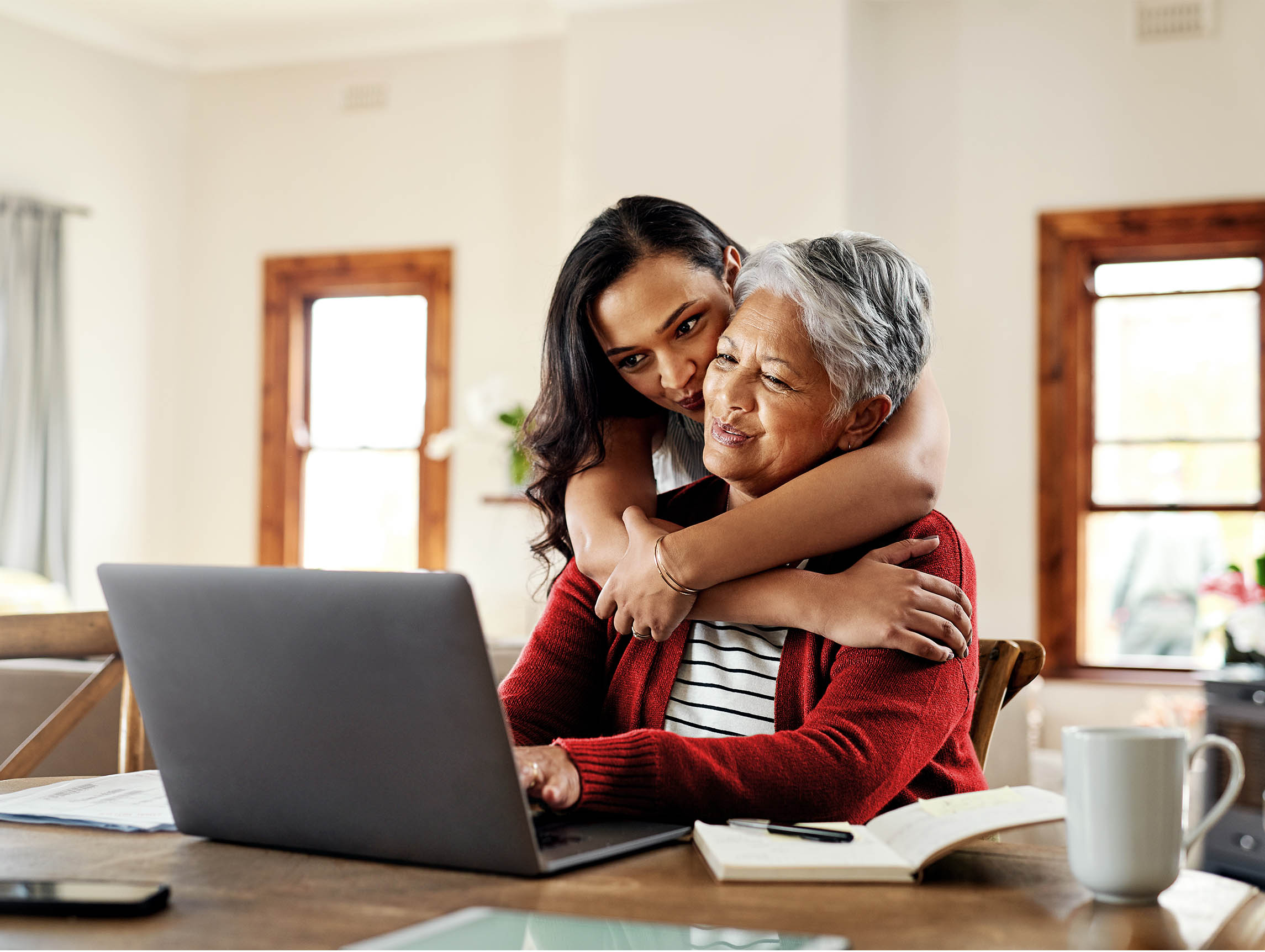 A woman hugging her mother from behind while her mother works on a laptop