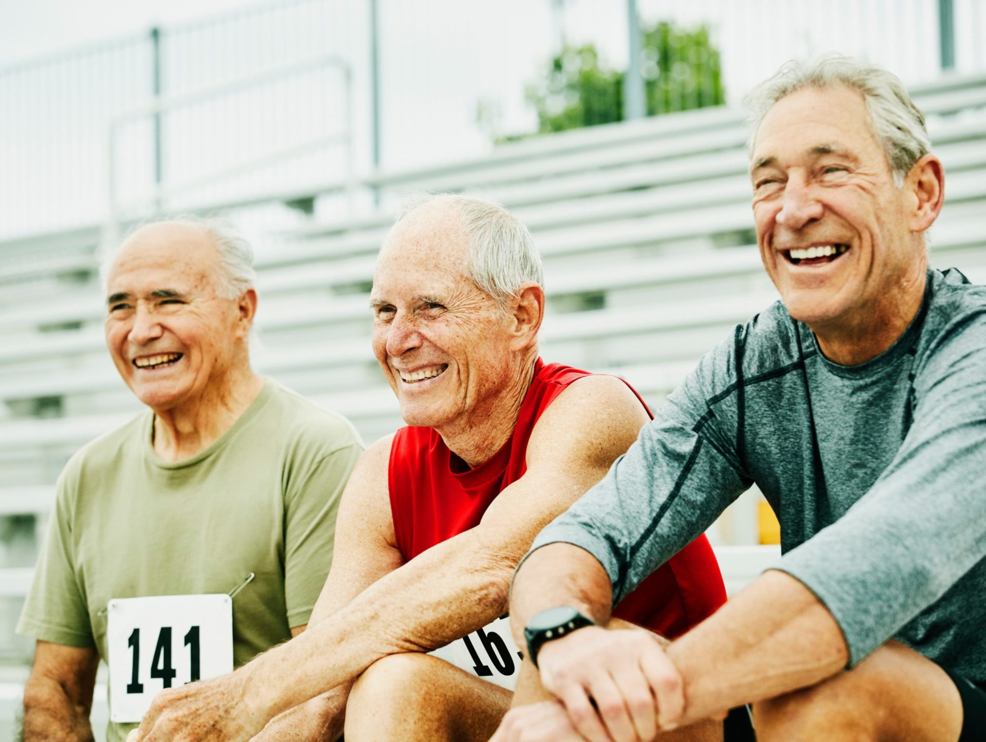 Three senior men sit on the bleachers wearing race numbers on their shirts